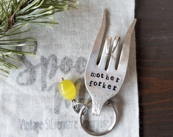 May include: A silver fork keychain with the words "mother forker" stamped on it. A yellow bead is attached to the keychain.