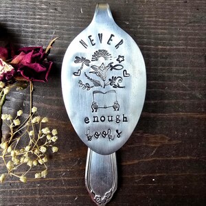 May include: A stamped metal spoon with the words "NEVER enough books" and floral illustrations. The spoon is silver-colored and rests on a dark wooden surface, with dried flowers in the background.