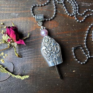 May include: A silver pendant necklace with a floral design. The pendant is shaped like a spoon and has a pink bead. The necklace is on a dark wooden surface with a dried rose and sprigs of dried flowers.