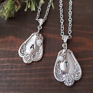 May include: Two silver necklaces with spoon pendants. The pendants are engraved with the letters "A" and "T" and have a small white bead attached to each.
