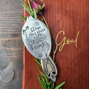 May include: A vintage silver spoon with the words "Chase your goals with grace & strength" stamped on it, resting on a red leather journal with the word "Goal" in gold script. The spoon is adorned with floral designs and fresh rosemary.