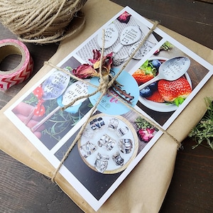 Pu&ograve; includere: Un pacco regalo avvolto in carta marrone e legato con spago. Il pacco contiene biglietti stampati con immagini di cucchiai, fragole e mirtilli. Sono visibili anche nastro adesivo decorativo e un gomitolo di spago.