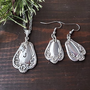 May include: Silver necklace and earrings set with a spoon pendant featuring the letter J and floral designs.