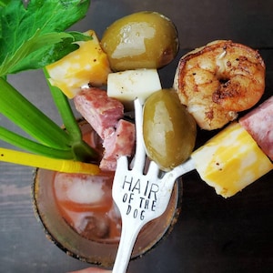 May include: A silver fork with the words "Hair of the Dog" engraved on the handle. The fork is holding a Bloody Mary garnish with cheese cubes, olives, a shrimp, and a slice of salami.