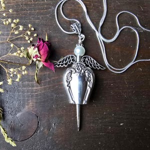 Pu&ograve; includere: Una collana con pendente argentato con un design ad ali d'angelo. Il pendente ha una perla madreperlacea e una base lunga e appuntita. Una rosa secca e rametti di fiori secchi sono accanto alla collana su una superficie di legno scuro.