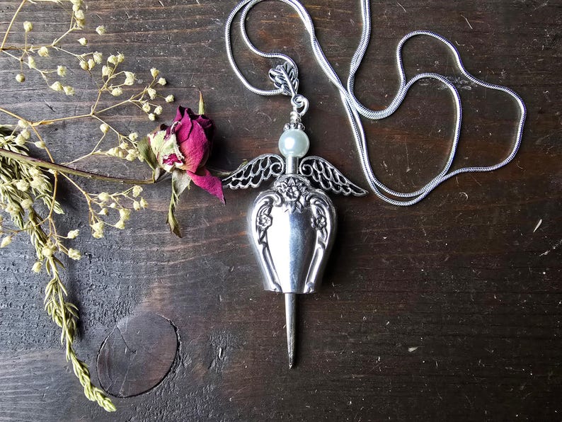 Pu&ograve; includere: Una collana con pendente argentato con un design ad ali d'angelo. Il pendente ha una perla madreperlacea e una base lunga e appuntita. Una rosa secca e rametti di fiori secchi sono accanto alla collana su una superficie di legno scuro.