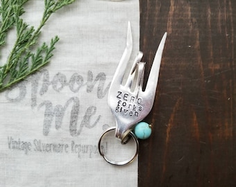 May include: A silver fork repurposed into a keychain with a turquoise bead. The fork has the words "Zero Forks Given" stamped on it.