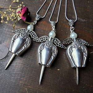 Pu&ograve; includere: Tre collane con ciondoli a forma di angelo in argento. Ogni ciondolo presenta un corpo a forma di tulipano, ali delicate e una perla. Le collane sono esposte su una superficie di legno scuro, con fiori secchi e una rosa sullo sfondo.