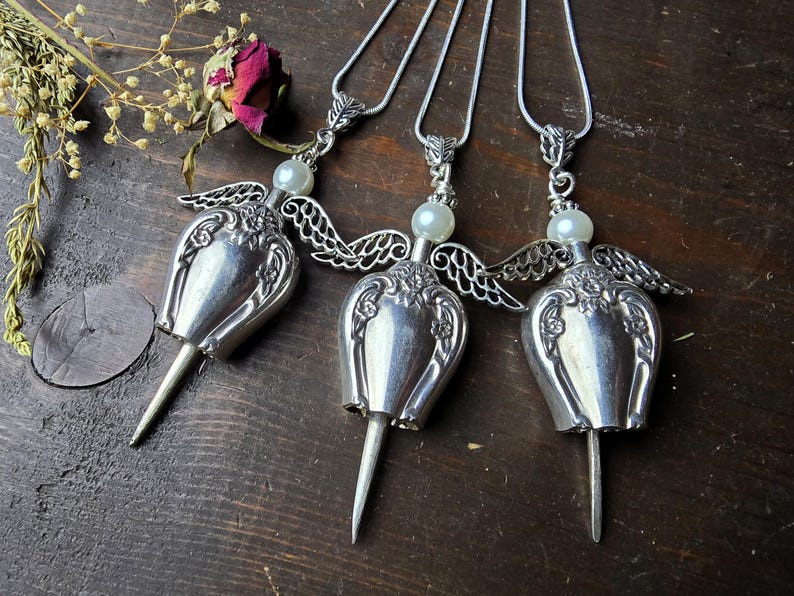 Pu&ograve; includere: Tre collane con ciondoli a forma di angelo in argento. Ogni ciondolo presenta un corpo a forma di tulipano, ali delicate e una perla. Le collane sono esposte su una superficie di legno scuro, con fiori secchi e una rosa sullo sfondo.
