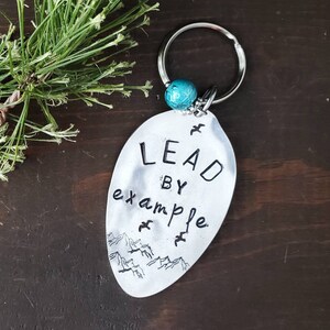 May include: Silver spoon keychain with a turquoise bead and the words "Lead by example" engraved on it. The spoon has a design of mountains and birds.