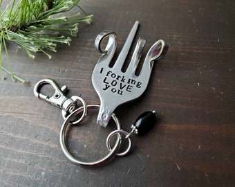 May include: A silver fork keychain with the words "I forking love you" engraved on it. The fork is bent into a heart shape and has a black bead charm attached to it.
