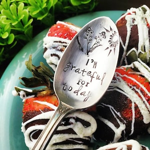 May include: A silver spoon with the words "I'm grateful for today" engraved on it. The spoon is resting on top of chocolate-covered strawberries with white chocolate drizzle.