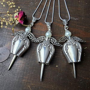 Pu&ograve; includere: Tre collane con ciondolo a forma di angelo in argento. Ogni collana presenta un corpo a forma di cucchiaio d'argento, ali delicate e una perla. I ciondoli sono sospesi a una catena d'argento, creando un gioiello unico e artistico.