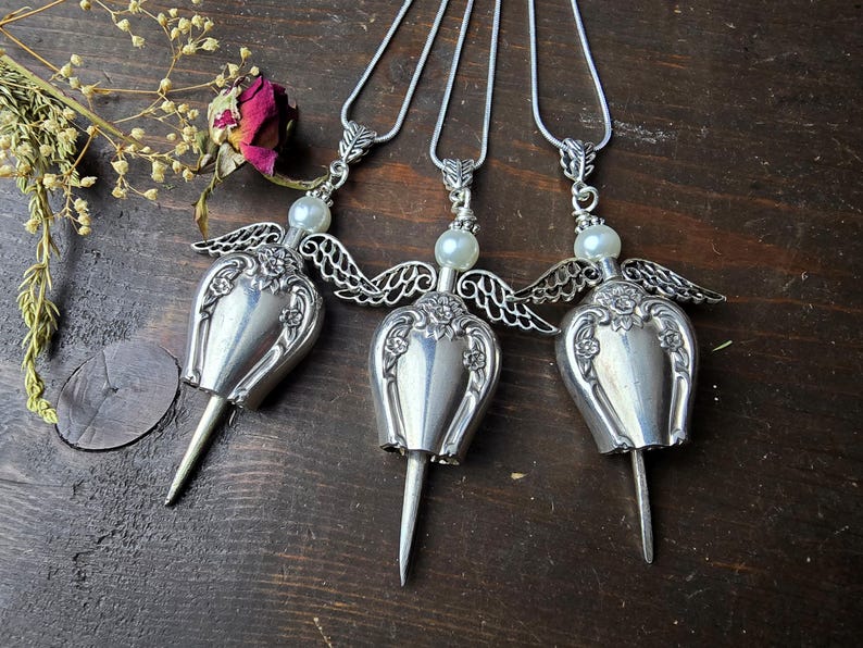 Pu&ograve; includere: Tre collane con ciondolo a forma di angelo in argento. Ogni collana presenta un corpo a forma di cucchiaio d'argento, ali delicate e una perla. I ciondoli sono sospesi a una catena d'argento, creando un gioiello unico e artistico.