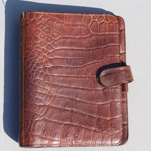 May include: A brown leather planner with a crocodile embossed pattern and a flap closure.