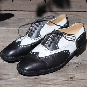 Op de afbeelding: Een paar tweekleurige zwarte en witte leren Oxford-schoenen met decoratieve perforaties. De schoenen hebben zwarte veters en een klassiek wingtip-ontwerp, geplaatst op een houten oppervlak.