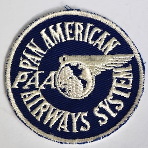 May include: A round, embroidered patch with a navy blue background and white text. The text reads "PAN AMERICAN AIRWAYS SYSTEM" and "PAA". A globe with wings is in the center. The patch has a white border.