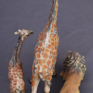 K&ouml;nnte beinhalten: Drei Vintage-Tierfiguren: zwei Giraffen und ein L&ouml;we. Die Giraffen haben orange-braune Fleckenmuster, w&auml;hrend der L&ouml;we hellbraun mit einer dunklen M&auml;hne ist. Die Figuren scheinen aus einem verwitterten Material zu bestehen.
