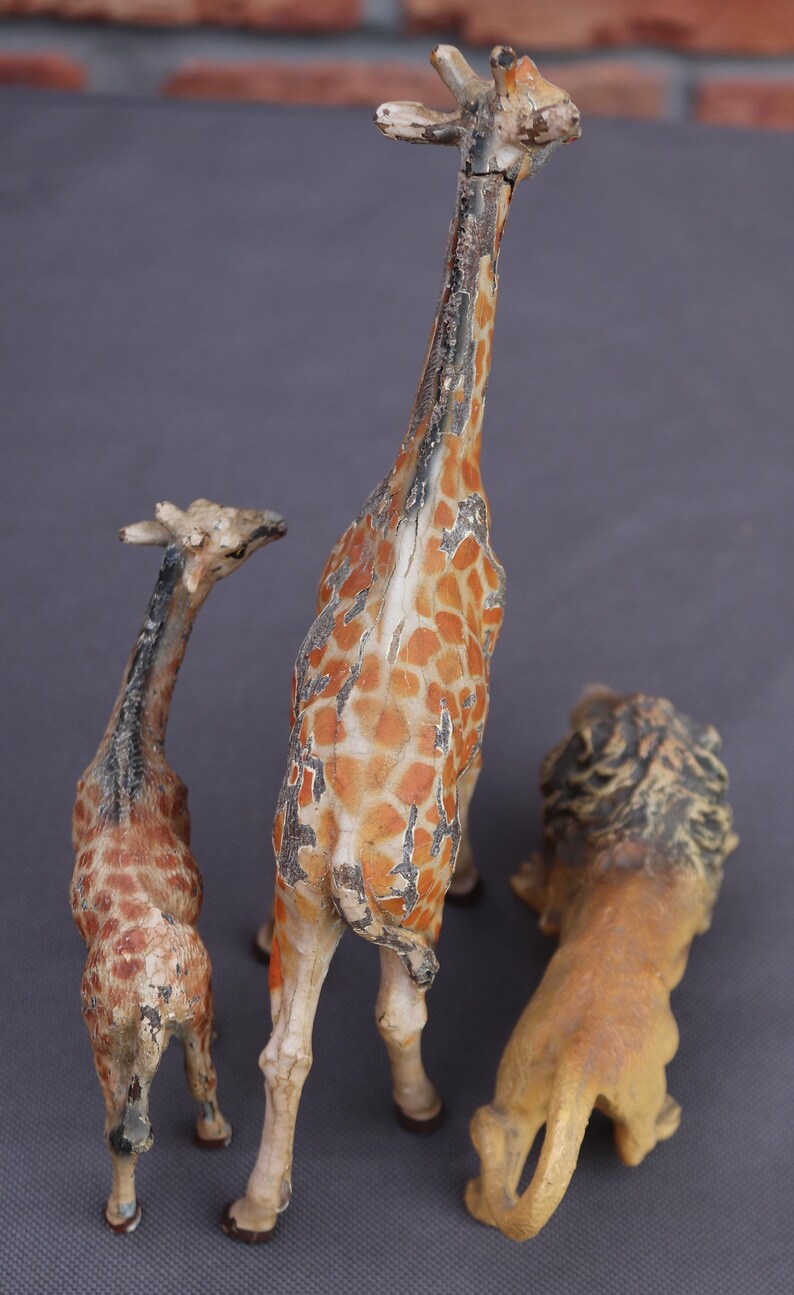 K&ouml;nnte beinhalten: Drei Vintage-Tierfiguren: zwei Giraffen und ein L&ouml;we. Die Giraffen haben orange-braune Fleckenmuster, w&auml;hrend der L&ouml;we hellbraun mit einer dunklen M&auml;hne ist. Die Figuren scheinen aus einem verwitterten Material zu bestehen.