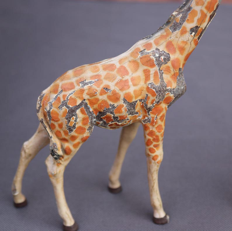 K&ouml;nnte beinhalten: Eine Vintage-Giraffenfigur mit einem Used-Look. Der K&ouml;rper der Giraffe ist beige mit orangefarbenen und schwarzen Flecken. Die Beine sind hellbeige und die Hufe sind dunkelbraun. Die Figur steht auf einer grauen Oberfl&auml;che.