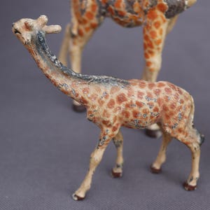 K&ouml;nnte beinhalten: Zwei antike Giraffenfiguren. Die kleinere Giraffe befindet sich im Vordergrund, mit einem langen Hals und einem gefleckten Muster in Braun- und Beiget&ouml;nen. Die gr&ouml;&szlig;ere Giraffe ist teilweise im Hintergrund zu sehen. Die Figuren haben ein antikes Finish.