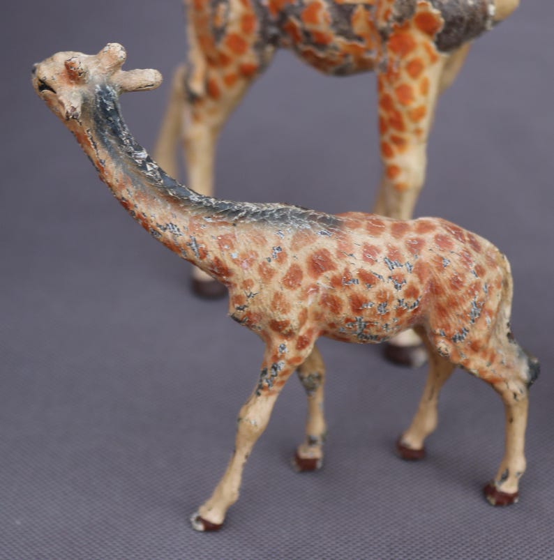 K&ouml;nnte beinhalten: Zwei antike Giraffenfiguren. Die kleinere Giraffe befindet sich im Vordergrund, mit einem langen Hals und einem gefleckten Muster in Braun- und Beiget&ouml;nen. Die gr&ouml;&szlig;ere Giraffe ist teilweise im Hintergrund zu sehen. Die Figuren haben ein antikes Finish.
