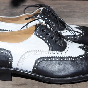 Op de afbeelding: Een paar tweekleurige leren Oxford-schoenen. De schoenen hebben een klassiek wingtip-ontwerp met zwarte en witte panelen, decoratieve perforaties en zwarte veters. De zolen zijn zwart.