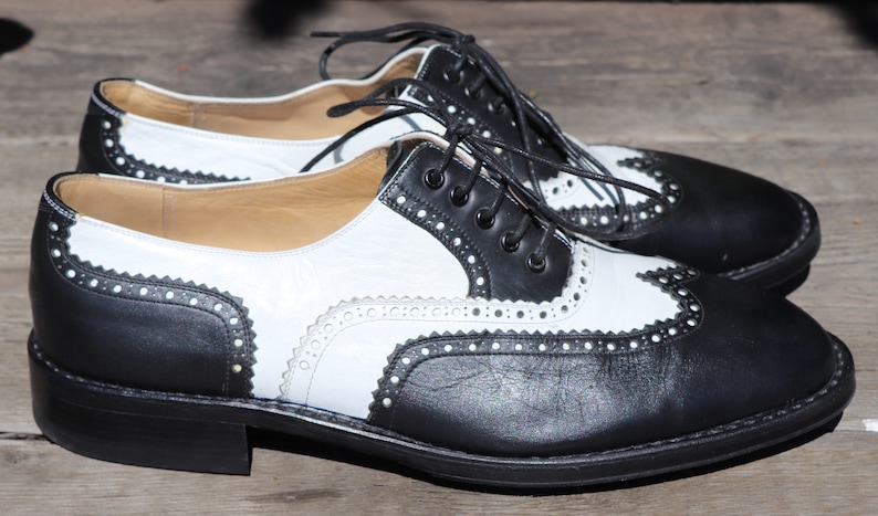 Op de afbeelding: Een paar tweekleurige leren Oxford-schoenen. De schoenen hebben een klassiek wingtip-ontwerp met zwarte en witte panelen, decoratieve perforaties en zwarte veters. De zolen zijn zwart.