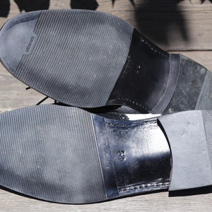 Op de afbeelding: Close-up van de zolen van twee zwart-witte leren herenschoenen. De zolen zijn gemaakt van zwart rubber met een textuurpatroon en de merknaam "Vibram" is zichtbaar. De schoenen staan op een houten ondergrond.
