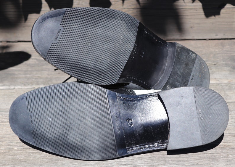 Op de afbeelding: Close-up van de zolen van twee zwart-witte leren herenschoenen. De zolen zijn gemaakt van zwart rubber met een textuurpatroon en de merknaam "Vibram" is zichtbaar. De schoenen staan op een houten ondergrond.