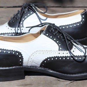 Op de afbeelding: Een paar tweekleurige wingtip schoenen. De schoenen zijn zwart en wit met decoratieve perforaties langs de randen. De veters zijn zwart en de schoenen staan op een houten oppervlak.