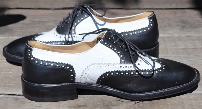 Op de afbeelding: Een paar tweekleurige wingtip schoenen. De schoenen zijn zwart en wit met decoratieve perforaties langs de randen. De veters zijn zwart en de schoenen staan op een houten oppervlak.