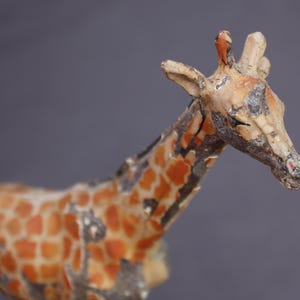 K&ouml;nnte beinhalten: Eine Vintage-Giraffenfigur mit verwittertem Aussehen. Die Giraffe hat einen beigefarbenen K&ouml;rper mit orangefarbenen und braunen Flecken. Kopf und Hals sind detailliert mit sichtbaren Abnutzungserscheinungen, die auf Alter und Gebrauch hindeuten. Der Hintergrund ist einfarbig grau.