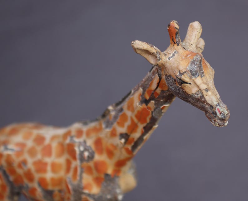 K&ouml;nnte beinhalten: Eine Vintage-Giraffenfigur mit verwittertem Aussehen. Die Giraffe hat einen beigefarbenen K&ouml;rper mit orangefarbenen und braunen Flecken. Kopf und Hals sind detailliert mit sichtbaren Abnutzungserscheinungen, die auf Alter und Gebrauch hindeuten. Der Hintergrund ist einfarbig grau.