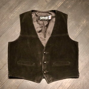 Op de afbeelding: Een zwart suède vest met knoopsluiting en twee zakken. Het vest heeft een label met de tekst "Schott NYC".