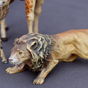 K&ouml;nnte beinhalten: Detaillierte Figur eines L&ouml;wen in geduckter Position, mit einem beigefarbenen K&ouml;rper und einer dunklen M&auml;hne. Das Maul des L&ouml;wen ist ge&ouml;ffnet und zeigt seine Z&auml;hne. Andere Tierfiguren, darunter eine Giraffe und ein Hirsch, sind im Hintergrund teilweise sichtbar.