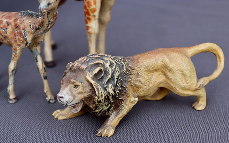 K&ouml;nnte beinhalten: Detaillierte Figur eines L&ouml;wen in geduckter Position, mit einem beigefarbenen K&ouml;rper und einer dunklen M&auml;hne. Das Maul des L&ouml;wen ist ge&ouml;ffnet und zeigt seine Z&auml;hne. Andere Tierfiguren, darunter eine Giraffe und ein Hirsch, sind im Hintergrund teilweise sichtbar.