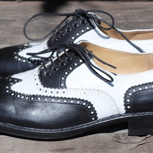 Op de afbeelding: Een paar tweekleurige wingtip-schoenen. De schoenen zijn van zwart en wit leer met decoratieve perforaties en stiksels. De veters zijn zwart en de zolen zijn zwart.