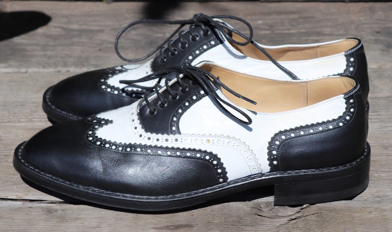 Op de afbeelding: Een paar tweekleurige wingtip-schoenen. De schoenen zijn van zwart en wit leer met decoratieve perforaties en stiksels. De veters zijn zwart en de zolen zijn zwart.