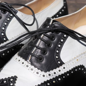 Op de afbeelding: Close-up van een paar tweekleurige Oxford-schoenen. De schoenen hebben een klassiek ontwerp met zwarte lederen accenten op de neus, hiel en vetersluiting, gecontrasteerd door witte lederen panelen. De schoenen hebben zwarte veters en decoratieve perforaties.