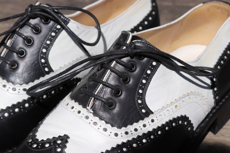 Op de afbeelding: Close-up van een paar tweekleurige Oxford-schoenen. De schoenen hebben een klassiek ontwerp met zwarte lederen accenten op de neus, hiel en vetersluiting, gecontrasteerd door witte lederen panelen. De schoenen hebben zwarte veters en decoratieve perforaties.