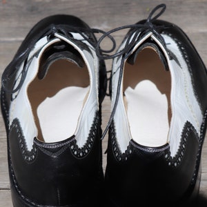 Op de afbeelding: Een paar tweekleurige Oxford-schoenen van zwart en wit leer. De schoenen hebben een klassiek wingtip-ontwerp met decoratieve perforaties langs de randen. Zwarte veters zijn bovenaan vastgebonden. De schoenen worden op een houten oppervlak weergegeven.