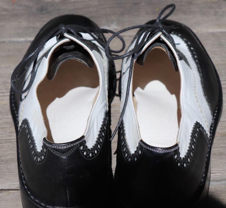 Op de afbeelding: Een paar tweekleurige Oxford-schoenen van zwart en wit leer. De schoenen hebben een klassiek wingtip-ontwerp met decoratieve perforaties langs de randen. Zwarte veters zijn bovenaan vastgebonden. De schoenen worden op een houten oppervlak weergegeven.