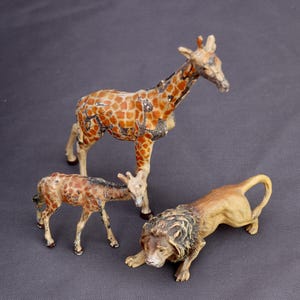 K&ouml;nnte beinhalten: Drei Vintage-Tierfiguren: eine Giraffe, eine Babygiraffe und ein L&ouml;we. Die Giraffe hat ein braun-beiges Fleckenmuster. Der L&ouml;we ist beige mit einer dunkelbraunen M&auml;hne. Die Figuren stehen auf einer grauen Oberfl&auml;che.