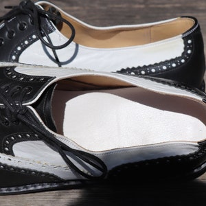 Op de afbeelding: Zwarte en witte leren Oxford-schoenen met decoratieve perforaties en zwarte veters. De schoenen hebben een klassiek wingtip-ontwerp met een wit bovenwerk en zwarte accenten. De zolen zijn lichtbruin.