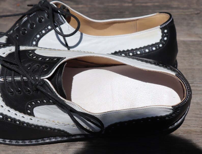 Op de afbeelding: Zwarte en witte leren Oxford-schoenen met decoratieve perforaties en zwarte veters. De schoenen hebben een klassiek wingtip-ontwerp met een wit bovenwerk en zwarte accenten. De zolen zijn lichtbruin.