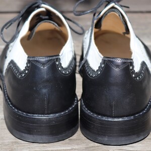 Op de afbeelding: Een paar tweekleurige leren Oxford-schoenen. De schoenen hebben een zwarte neus en hiel met een wit bovenwerk. Het ontwerp omvat decoratieve perforaties en zwarte veters. De zolen zijn zwart en hebben een dikke, gestapelde uitstraling.
