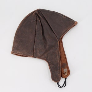 May include: A brown leather aviator hat with ear flaps and a chin strap. The hat has a distressed appearance, with creases and colour variations. The ear flaps extend downwards, and the chin strap is secured with a black cord.