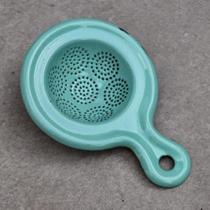 May include: A light teal tea strainer with a handle and a perforated bowl. The strainer has a circular bowl with a handle that has a hole at the end for hanging. The tea strainer is made of metal.