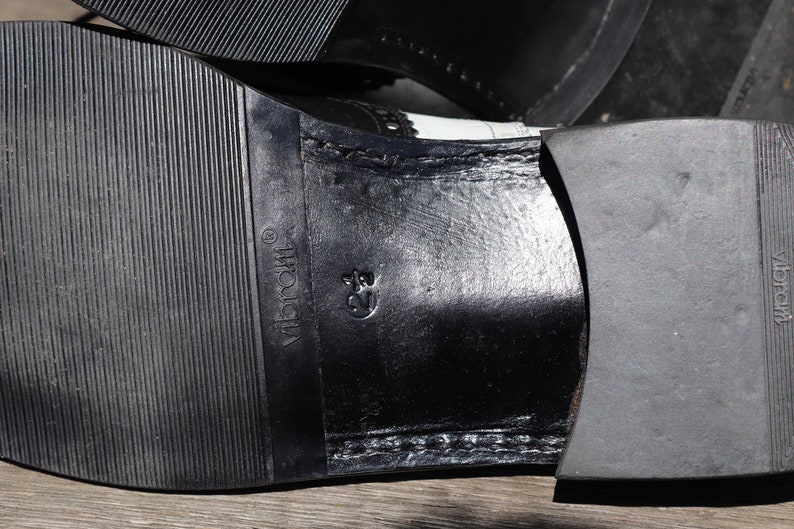 Op de afbeelding: Close-up van de zool van een zwart-witte leren schoen. De rubberen zool heeft een textuurpatroon met de merknaam "Vibram" zichtbaar. De schoenmaat is gemarkeerd als 39.